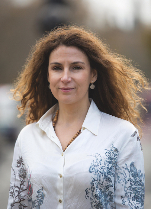 Pamela Dow, standing outside, wearing a white shirt with a blue design on it. Her long, curly brown hair blows out behind her